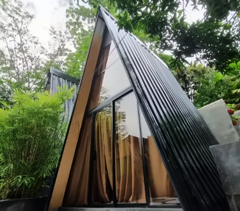 Exterior de una cabaña en A con techo de lámina negra y fachada de vidrio con cortinas, entre bambú y árboles.