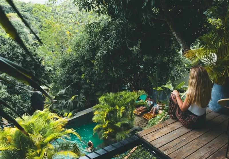 Una huésped sentada en una terraza de madera mira hacia la piscina, entre palmas, donde otras personas descansan en tumbonas; alrededor, árboles altos y selva.