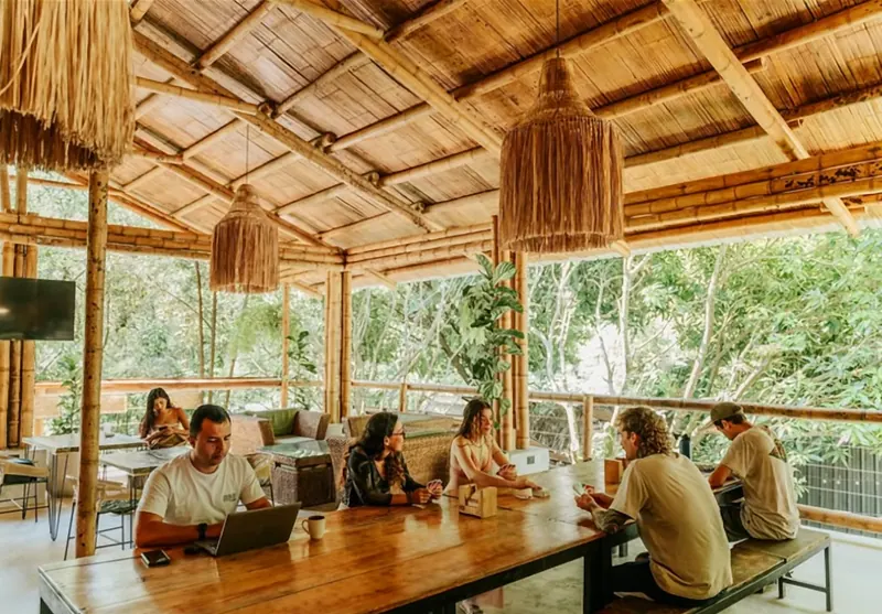 Zona social bajo un techo de bambú y caña: personas con portátiles y tazas conversan alrededor de una mesa larga de madera, con lámparas colgantes de fibra y árboles detrás de la baranda.