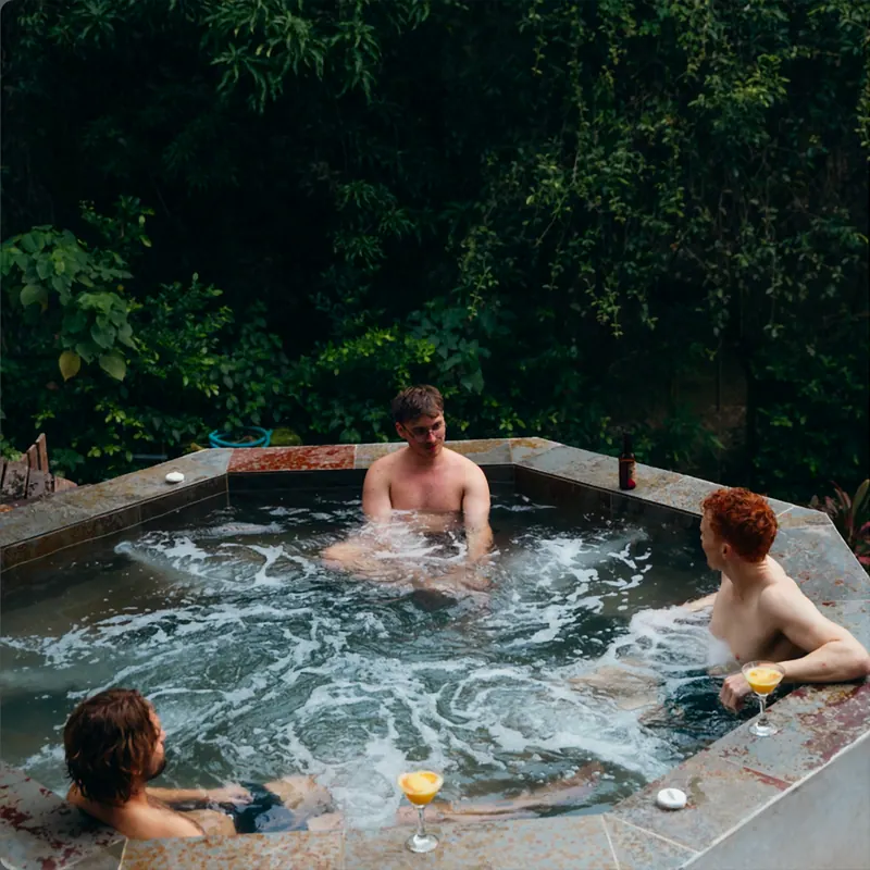 Tres amigos conversan en el jacuzzi de borde de piedra mientras el agua burbujea; hay dos cócteles sobre el borde y una pared de vegetación detrás.