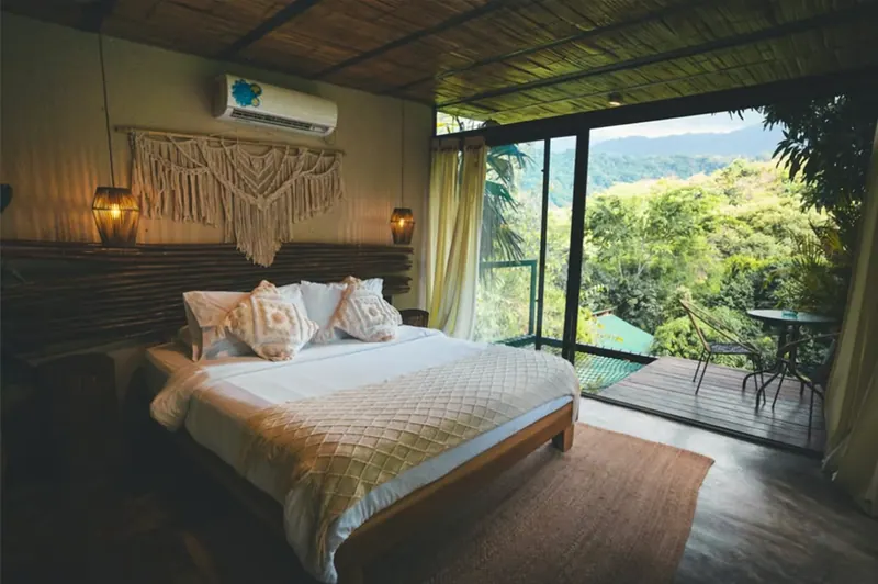 Habitación Montaña: una cama amplia con cojines tejidos, macramé sobre la cabecera y lámparas de fibra, frente a un ventanal de piso a techo abierto a un balcón de madera con vista a la selva y las montañas.