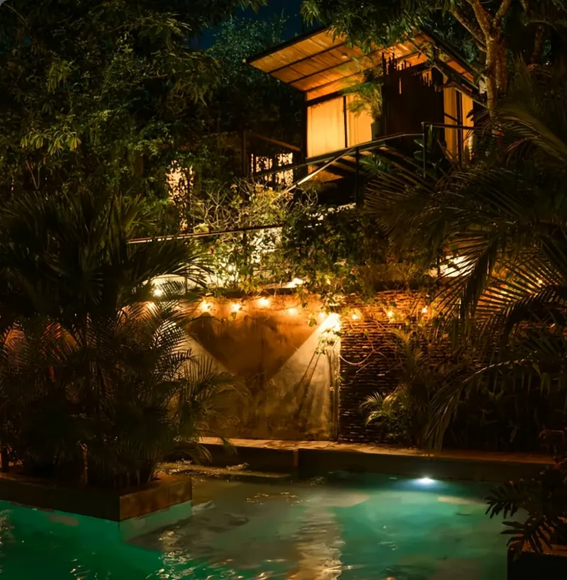La piscina de noche, iluminada en verde agua, con una guirnalda de luces sobre un muro de piedra y una cabaña elevada con luz cálida entre las palmas.