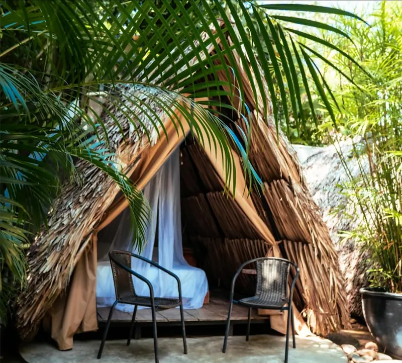 Cabaña en forma de tipi con techo de palma tejida; la entrada tiene un tul blanco y deja ver una cama, con dos sillas negras al frente y palmas alrededor.