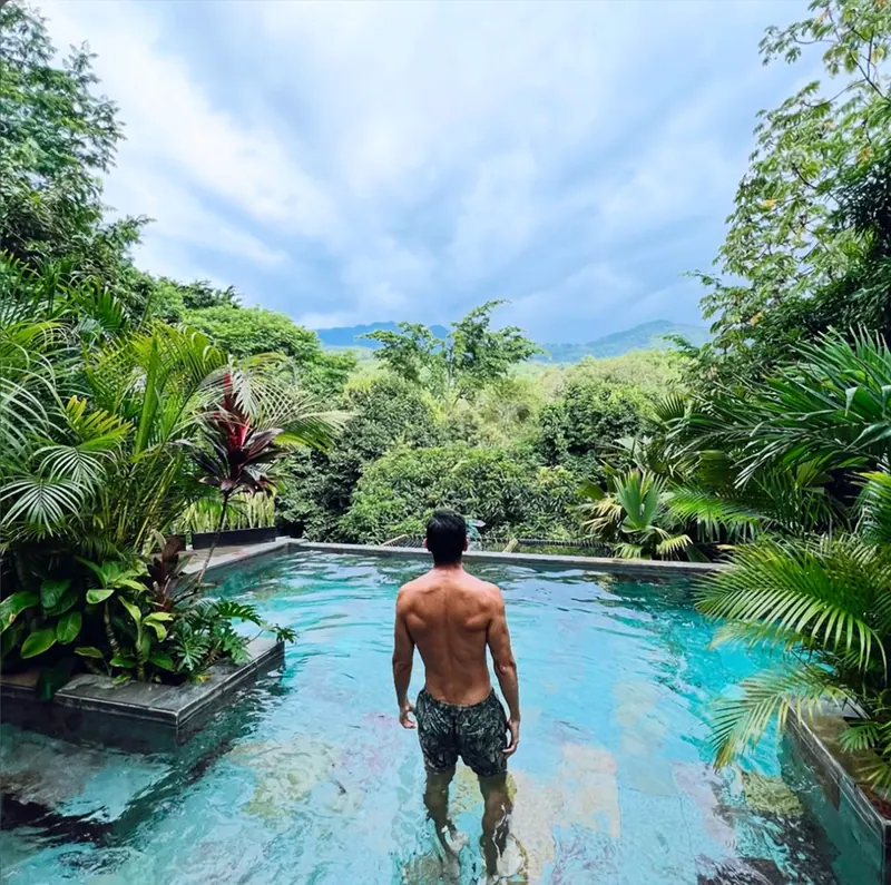 Un huésped de espaldas, con el agua a las rodillas, en la piscina rodeada de palmas y plantas tropicales; al fondo, bosque y montañas bajo un cielo nublado.