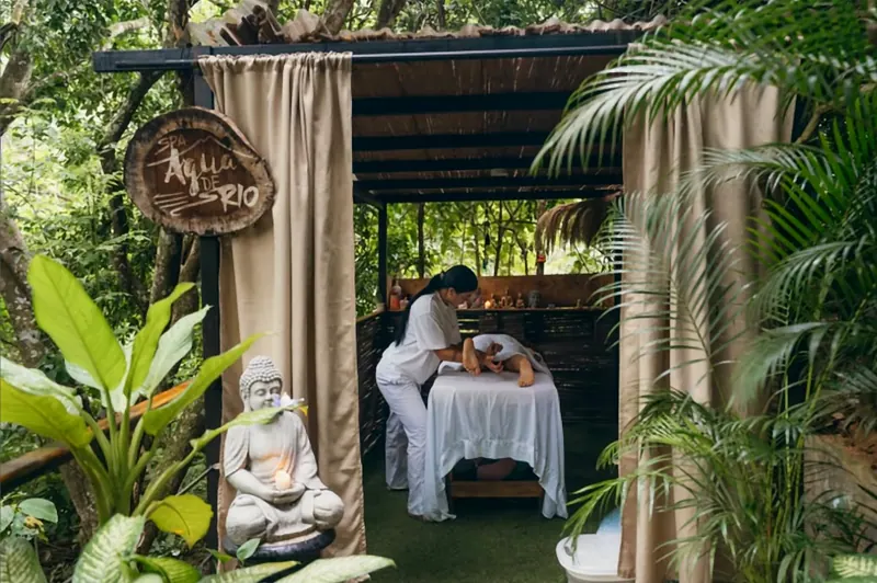Espacio de masajes entre la vegetación: una terapeuta atiende a una persona en una camilla bajo un toldo con cortinas, junto al letrero tallado «Agua de Río» y una figura de Buda con una vela.