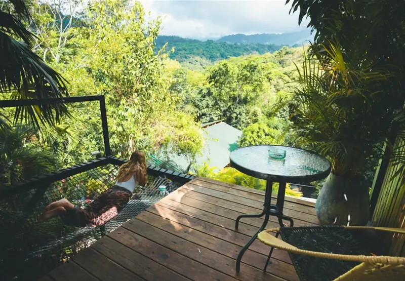 Terraza de madera con una malla tendida sobre el vacío, donde descansa una huésped; al lado, una mesa redonda de vidrio y una silla tejida, y al frente, selva y montañas.